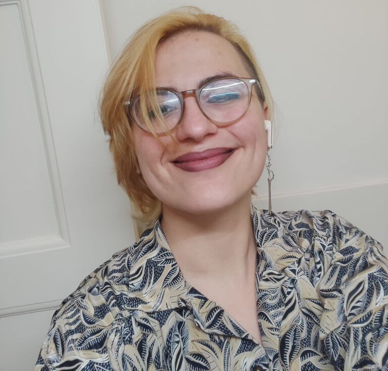 Porträt von Annamaria Balov, einer blonden Frau mit Brille und blumigem Shirt, die direkt in die Kamera blickt.